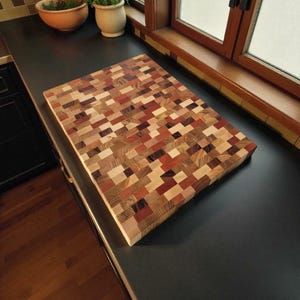 Puede incluir: Una tabla de cortar rectangular de testa, hecha de varias tonalidades de madera, incluyendo marrones claros, medios y oscuros, y tonos rojizos. La tabla está compuesta por pequeños bloques rectangulares dispuestos en un patrón de mosaico. La tabla mide unos 46 cm de largo y 30 cm de ancho.