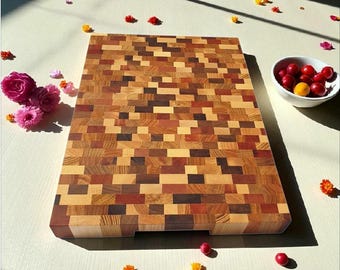 May include: A rectangular end-grain cutting board made of various wood tones, including light and dark browns, and reds. The board has a handle cutout on one side. A small white bowl filled with red and yellow fruit sits nearby.