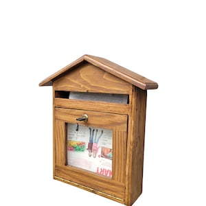 May include: A wooden mailbox with a pitched roof and a clear window displaying a newspaper. The mailbox is stained a warm brown color and has a small key lock. The design is reminiscent of a small house.