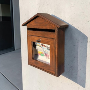 May include: A wooden mailbox with a pitched roof, mounted on a concrete wall. The mailbox is stained a warm brown color and has a glass window on the front, revealing colorful contents. A key is in the lock.