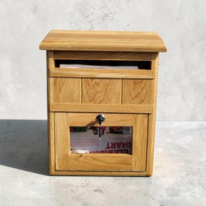 Puede incluir: Un buzón de madera con techo inclinado y puerta delantera con cerradura. El buzón, de madera clara, tiene una ranura para el correo en la parte superior. La puerta tiene una ventana transparente que muestra un periódico con la palabra "SMART".