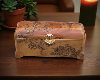 Puede incluir: Caja de joyería de madera con un diseño decorativo de ramas de árboles. La caja es rectangular con una parte superior curvada y un cierre dorado. La madera tiene una veta natural y un tono marrón rojizo. Una vela encendida y una planta en maceta están en el fondo.