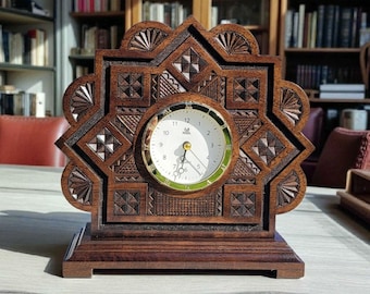 May include: A wooden desk clock with an intricate, geometric carved design. The clock face is white with gold-colored hands and a green border. The clock sits on a rectangular base and has a starburst-like frame. The wood has a rich, dark brown color.