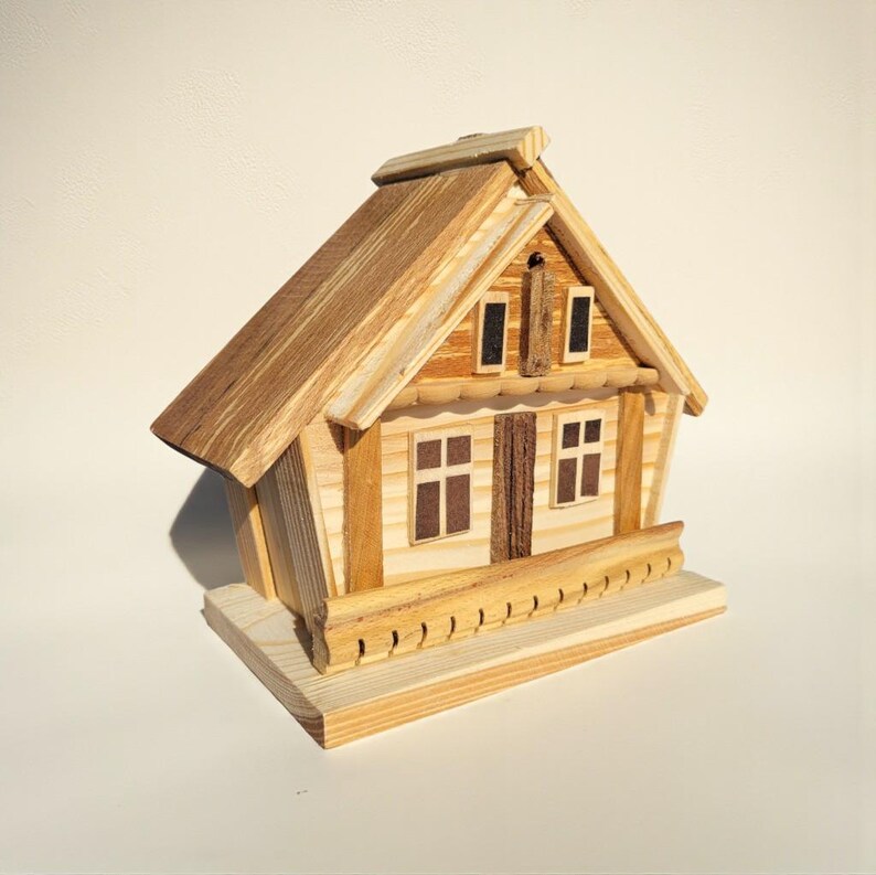 Puede incluir: Una casa de madera en miniatura con tejado y paredes de color marr&oacute;n claro. La casa tiene dos ventanas, una puerta y una peque&ntilde;a chimenea. Una barandilla decorativa recorre la parte delantera. La casa est&aacute; hecha de madera de varios tonos.