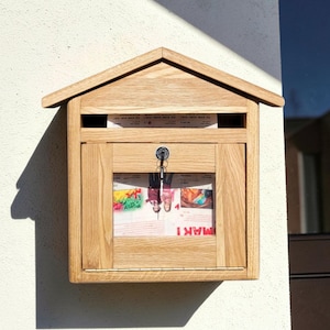 May include: A wall-mounted wooden mailbox with a gabled roof design, featuring a natural light brown wood grain finish. The front panel includes a rectangular glass window, a metal key lock with a silver key inserted, and a top mail slot opening.