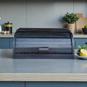 Könnte beinhalten: Eine dunkelgraue Brotdose aus Holz mit Rolldeckel und kleinem rechteckigem Griff. Die Box besteht aus Holz mit sichtbarer Maserung und steht auf einer hellgrauen Arbeitsplatte. Die Küchenszene umfasst eine Pflanze, Schneidebretter und eine Zitrone.