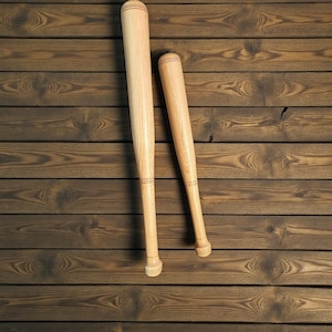 May include: Two wooden baseball bats, one full-sized and one smaller, rest on a textured, brown wooden surface. The bats are light-colored with a smooth finish and rounded ends, showcasing the natural wood grain.