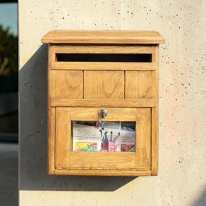 Op de afbeelding: Een houten brievenbus met een natuurlijke afwerking, gemonteerd op een getextureerde muur. De brievenbus heeft een gleuf voor post aan de bovenkant en een deur met een sleutelslot. Een krant is zichtbaar door het glazen raam. De brievenbus is rechthoekig met een schuine bovenkant.