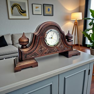 Pode incluir: Um relógio de lareira de madeira marrom escuro com um mostrador branco e entalhes ornamentados. O relógio tem um topo e uma base decorativos, com dois pináculos em cada lado. O relógio está sobre uma superfície cinza.