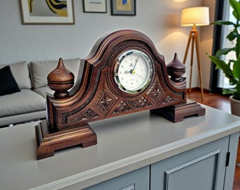 Può includere: Un orologio da mensola in legno marrone scuro con un quadrante bianco e intagli ornati. L'orologio ha una parte superiore e una base decorative, con due pinnacoli su entrambi i lati. L'orologio è posto su una superficie grigia.