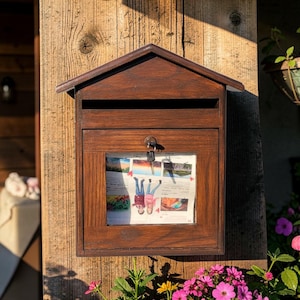 May include: A wooden mailbox with a house-shaped roof, stained a dark brown. The front features a framed window displaying a colorful image. A key hangs in the lock. The mailbox is mounted on a wooden post, surrounded by flowers.