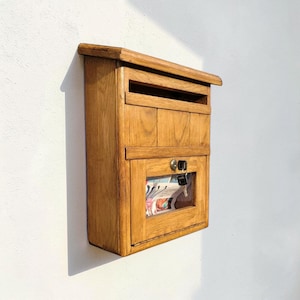 Peut inclure: Une boîte aux lettres en bois avec une finition naturelle est fixée sur un mur blanc. La boîte aux lettres a une fente pour le courrier et un compartiment inférieur avec une fenêtre en verre, révélant des journaux. Une clé est dans la serrure.