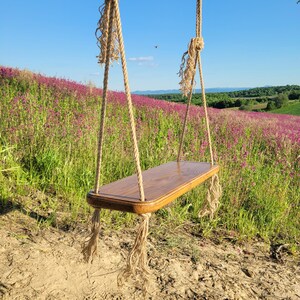 Peut inclure: Une balançoire en bois avec une assise rectangulaire et des bords arrondis est suspendue à une épaisse corde nouée. La balançoire est placée devant un champ de fleurs violettes et un ciel bleu clair.