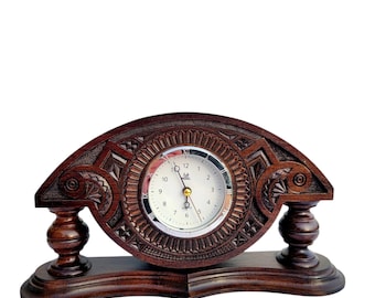 Puede incluir: Un reloj de madera marrón oscuro con intrincados tallados. La esfera del reloj es blanca con números y manecillas negros. El reloj está sobre una base de madera.