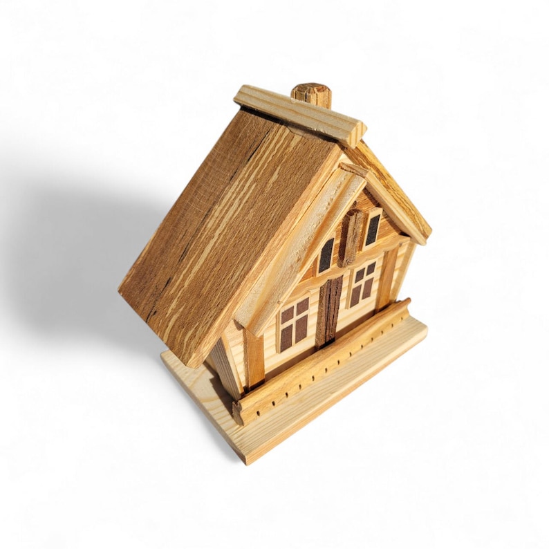 Puede incluir: Una casa de madera en miniatura con tejado a dos aguas, ventanas y chimenea. La casa est&aacute; construida con madera marr&oacute;n clara y oscura, con vetas visibles. Se asienta sobre una base rectangular.