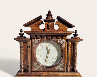 May include: A wooden mantel clock with a light-colored clock face and gold-colored hands. The clock has a dark brown wooden frame with decorative carvings and ornate metal accents. The clock is a classic, vintage style.