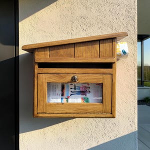 Può includere: Una cassetta delle lettere in legno con tetto spiovente è montata su un muro strutturato. La cassetta delle lettere ha una finestra di vetro che mostra la posta e un piccolo rotolo di carta con un motivo giallo e blu è infilato di lato.