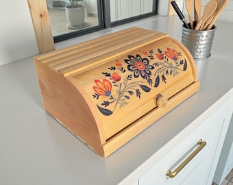 May include: A light-colored wooden bread box with a curved top and a floral design in orange, blue, and gray. The box has a wooden knob and a slatted top. The bread box is sitting on a white countertop.