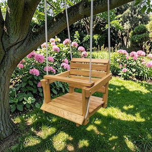 May include: A wooden bucket swing with a slatted seat, backrest, and front safety bar, suspended by two thick white ropes from a tree branch. The swing is constructed from light brown wood with visible grain and metal hardware at the joints.