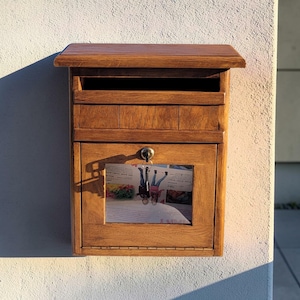 Könnte beinhalten: Ein wandmontierter Briefkasten aus Holz in mittelbrauner Ausführung mit einem oberen Einwurfschlitz, einer aufklappbaren Fronttür mit zentralem Schlüsselschloss und einem rechteckigen Glasfenster, das eine bedruckte Werbeanzeige zeigt.