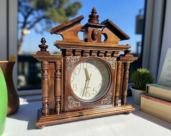 May include: A wooden antique clock with a light-colored clock face and gold hands. The clock has a decorative wooden frame with ornate details and a small roof-like structure on top. The clock is sitting on a white surface.