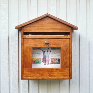 May include: A wooden mailbox with a pitched roof, stained in a warm brown hue. The mailbox has a key lock and a window displaying a newspaper. The mailbox is mounted on a white, vertical-slatted wall.