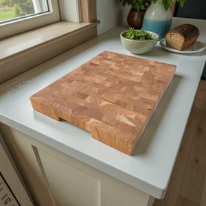 May include: A rectangular butcher block cutting board made of light brown wood. The end-grain construction is visible, showing a mosaic pattern. The board has a thick profile and is placed on a white countertop. It is a kitchen item.