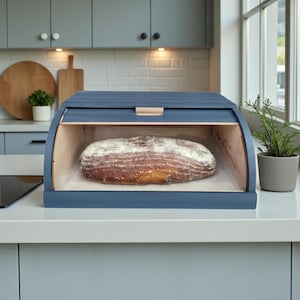 May include: A blue wooden bread bin with a roll-top lid, displaying a loaf of bread. The bread bin sits on a white countertop in a kitchen. The bread is approximately 30 cm long.