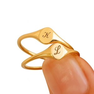Puede incluir: Dos anillos de sello en tono dorado pulido con caras lisas y redondeadas. Un anillo está grabado con la letra cursiva K y el otro anillo está grabado con la letra cursiva L. Los anillos tienen bandas delgadas y un acabado metálico brillante.