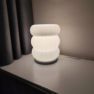May include: A white, ribbed, cylindrical table lamp with a soft glow. The lamp has a stacked design, with three rounded sections. The lamp is on a white surface, with a dark curtain in the background.