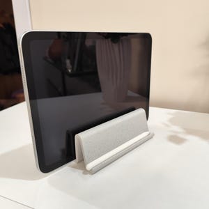 May include: A silver tablet rests in a speckled gray and white tablet stand. The stand has a curved design with a white accent. The tablet's screen is black, and the stand is on a white surface.