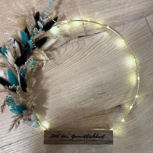 Peut inclure: Une couronne décorative illuminée avec une base en bois. La couronne est ornée de fleurs séchées de couleur teal, noire et beige. Le texte "Zeit für Gemütlichkeit" est gravé sur la base en bois. La couronne est éclairée par de petites lumières blanches chaudes.