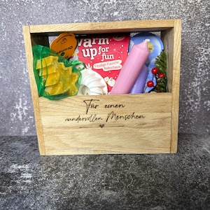 Könnte beinhalten: Eine Holzgeschenkbox gefüllt mit verschiedenen Artikeln. Darin befinden sich eine rosa Kerze, ein Buch mit dem Text "warm up for fun", ein grüner Netzbeutel mit gelben sternförmigen Gegenständen und weitere Dekorationen. Die Box trägt den Text "Für einen wundervollen Menschen".