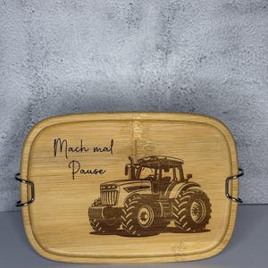 Könnte beinhalten: Eine rechteckige Bambus-Lunchbox mit einer Traktor-Gravur. Die Box hat eine hellbraune Farbe und trägt den Text "Mach mal Pause" in schwarzer Schrift. Metallverschlüsse sichern den Deckel.