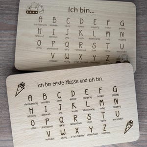 Può includere: Due lavagne alfabetiche in legno con lettere e parole incise. Una dice "Ich bin..." e l'altra "Ich bin erste Klasse und ich bin...". Ogni lettera ha una parola corrispondente in tedesco.