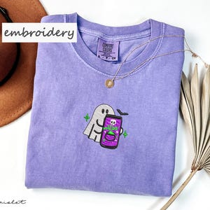 May include: Violet-colored Comfort Colors t-shirt with an embroidered ghost holding a phone with a potion design. The shirt has a crew neck and is accessorized with a gold necklace. The word "embroidery" is written on the top left of the image.