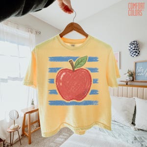 May include: A pale yellow short-sleeve t-shirt featuring a graphic of a red apple with a green leaf centred over horizontal blue brushstroke stripes. The shirt has a crew neckline and the Comfort Colors brand logo printed on the inner neck label.