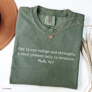 May include: Olive green Comfort Colors t-shirt with the text "God is our refuge and strength, a very present help in trouble. Psalm 46:1." A gold necklace with a pendant rests on the shirt. A brown hat is visible in the background.
