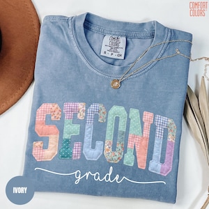 May include: A light blue Comfort Colors t-shirt with the word "SECOND" in colorful patchwork letters and the word "grade" in cursive. A gold necklace with a pendant is on the shirt. The shirt is size S-P-CH.