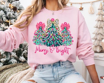 May include: Light pink sweatshirt featuring a festive Christmas tree design. The graphic includes three decorated trees with pink bows and the words "Merry & Bright" in a script font. The sweatshirt is a long-sleeved crew neck.
