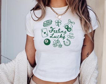 May include: White crop top with green graphic illustrations of lucky symbols, including a four-leaf clover, dice, and a horseshoe. The text "Feeling Lucky" is also printed on the shirt.