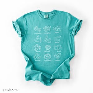 Op de afbeelding: Een turquoise t-shirt met een witte afbeelding van verschillende soorten pasta met hun namen eronder geschreven. De namen van de pasta zijn:  conchiglie, conchigliette, farfalle,  jamore, fusilli, spaghettini,  gnocchi,  bucatini, ravioli,  macaroni,  pappardelle,  orecchiette.