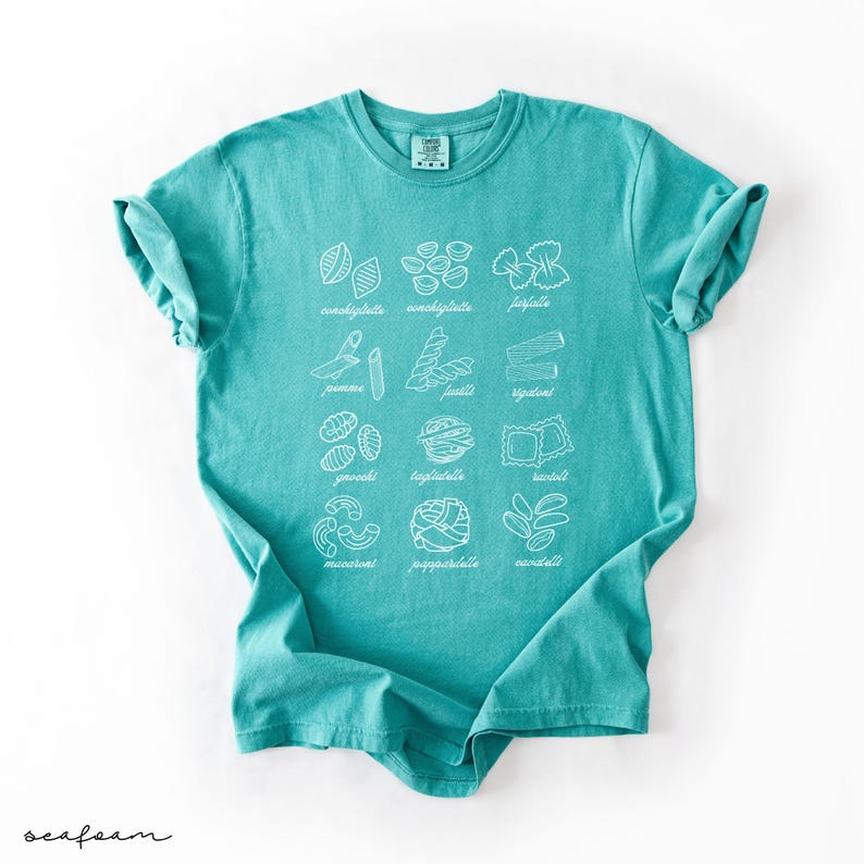 Op de afbeelding: Een turquoise t-shirt met een witte afbeelding van verschillende soorten pasta met hun namen eronder geschreven. De namen van de pasta zijn:  conchiglie, conchigliette, farfalle,  jamore, fusilli, spaghettini,  gnocchi,  bucatini, ravioli,  macaroni,  pappardelle,  orecchiette.