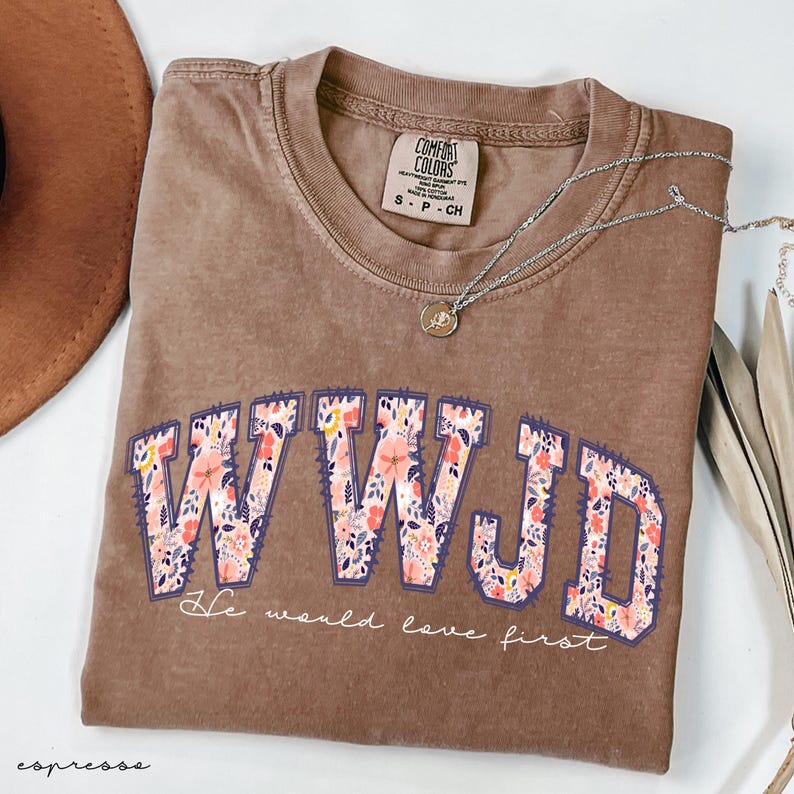 May include: Espresso-coloured t-shirt with the letters "WWJD" in a floral design. The phrase "He would love first" is below the letters. A silver necklace with a pendant is on the shirt.