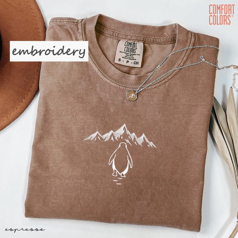 May include: Espresso-colored t-shirt with a white embroidered penguin and mountain design. The shirt features the text "Comfort Colors" and a small tag with size information. A silver necklace with a pendant is draped across the shirt.