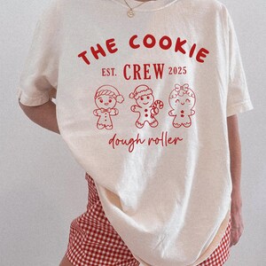 May include: Cream-colored t-shirt with the text "THE COOKIE CREW EST. 2025" in red. Below the text are three gingerbread people illustrations. The words "dough roller" are written below the gingerbread people in red.