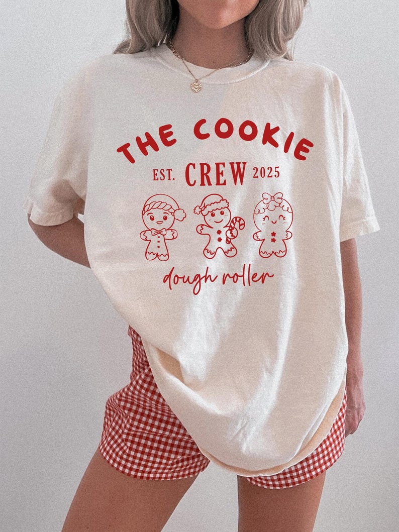 May include: Cream-colored t-shirt with the text "THE COOKIE CREW EST. 2025" in red. Below the text are three gingerbread people illustrations. The words "dough roller" are written below the gingerbread people in red.