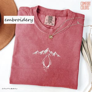 May include: Crimson-colored t-shirt with a white embroidered penguin and mountain design. The shirt has a crew neck and short sleeves. A silver necklace with a pendant is draped across the shirt. The word "embroidery" is in a white box.