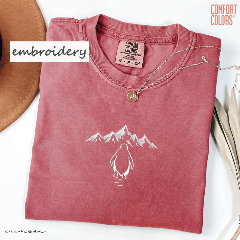 May include: Crimson-colored t-shirt with a white embroidered penguin and mountain design. The shirt has a crew neck and short sleeves. A silver necklace with a pendant is draped across the shirt. The word "embroidery" is in a white box.