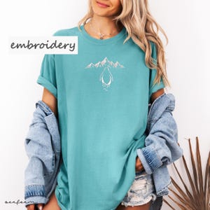 May include: Teal-colored t-shirt with a white embroidered design of a penguin in front of mountains. The word "embroidery" is printed in white on the upper left. The shirt has rolled-up sleeves.